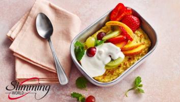 A small baking tin with oats, mixed fruit, yogurt, and mint leaves. A cloth napkin with a spoon on top is to the left of the baked oats.