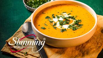 Carrot and coriander soup in a white bowl with herbs sprinkled on top. The bowl is on top of a brown cloth with a spoon to the left.