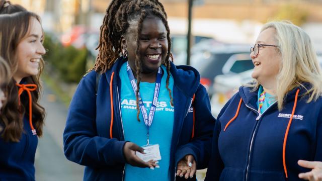 In this image, three Diabetes UK volunteers can be seen talking to each other. They are dressed in pale blue Diabetes UK t shirts and navy blue Diabetes UK hoodies.They are smiling and laughing. 