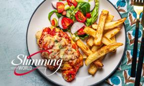 A chicken breast topped with tomato sauce, cheese, and herbs. A small side salad with tomatoes and sliced radishes and a small side of chips are on the side